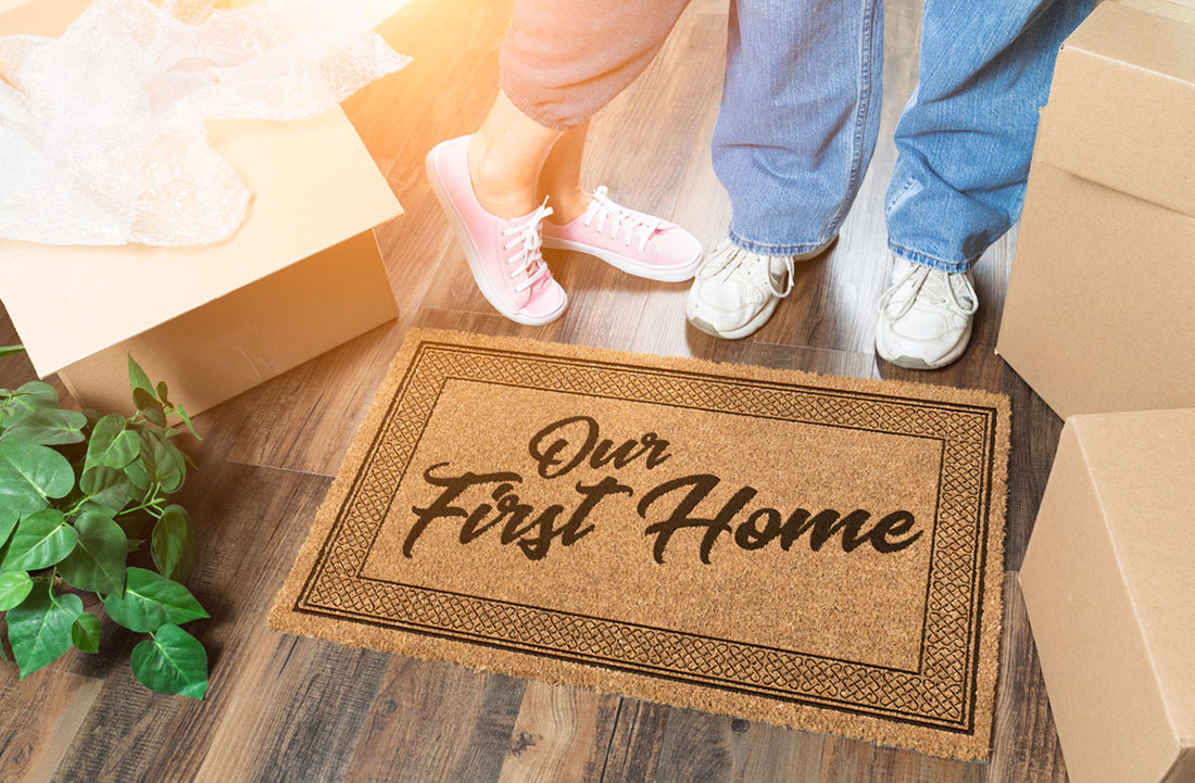 First-time homebuyers standing at the doorway of their new home with a welcome mat that says 'Our First Home'